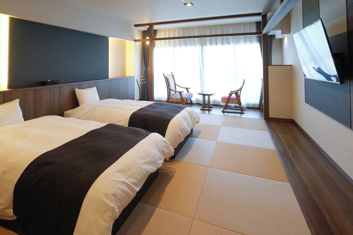 Une chambre d’hôtel avec deux lits, un coin salon avec une chaise et une table, et une grande fenêtre avec des rideaux.