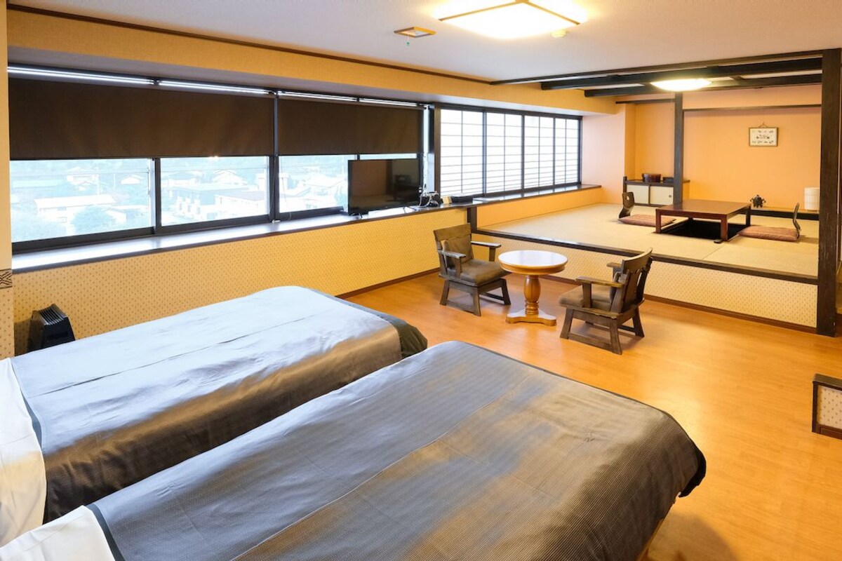 Habitación de hotel con dos camas, una mesita, sillas, televisor y un ventanal grande.