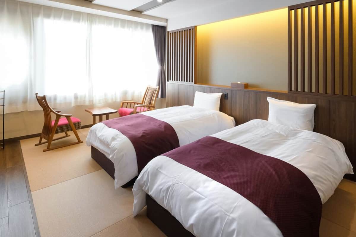Une chambre d’hôtel avec deux lits, un bureau, une chaise et une grande fenêtre avec des rideaux.