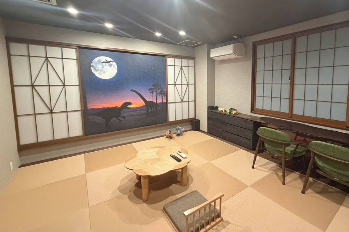 Une pièce de style japonais traditionnel, avec un sol recouvert de tatamis, une table basse, des chaises vertes, une grande fenêtre ornée de paravents shoji et une fresque représentant un dinosaure et la lune.