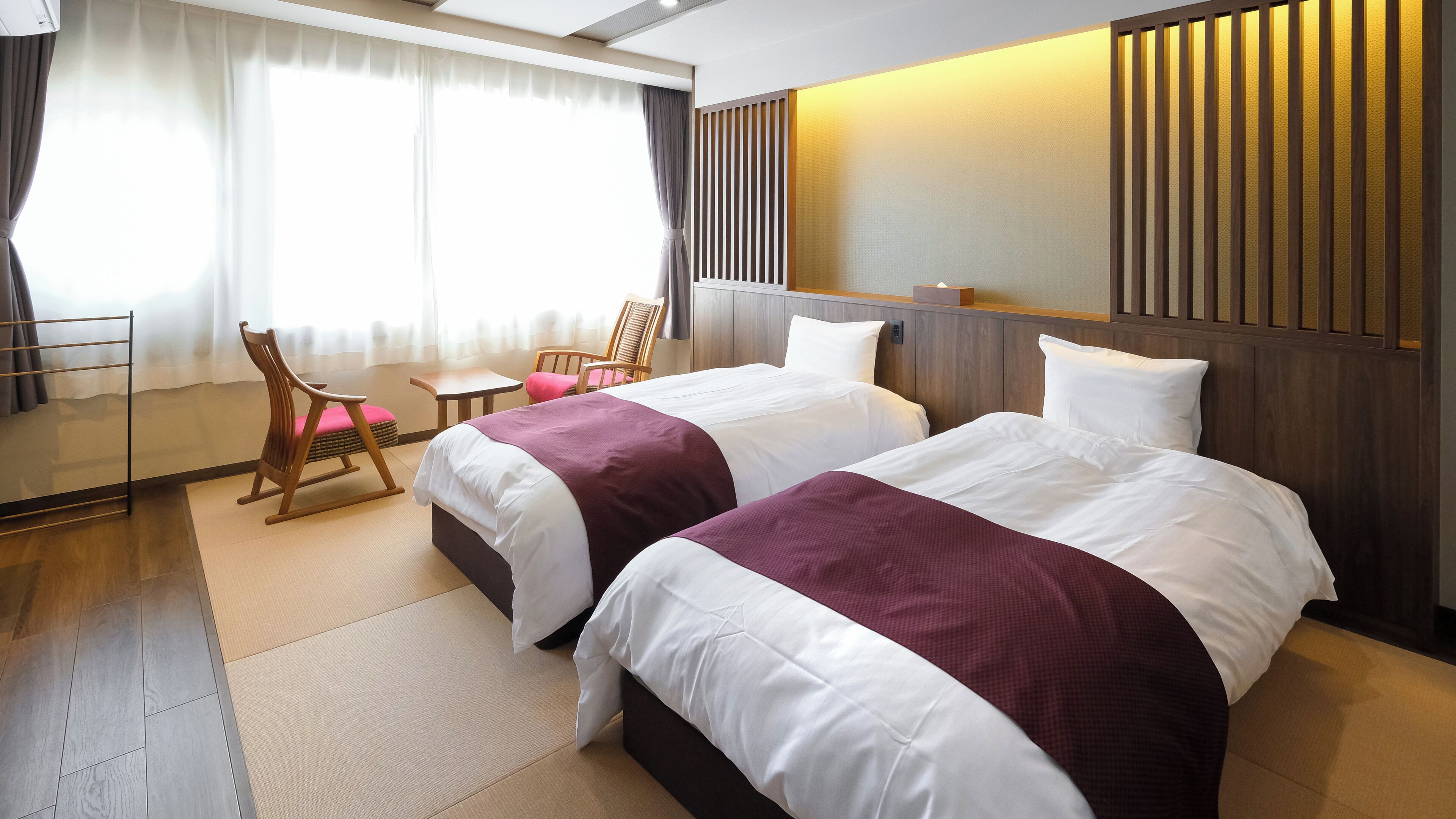 A hotel room with two beds, a small table, chairs, and a large window with curtains.