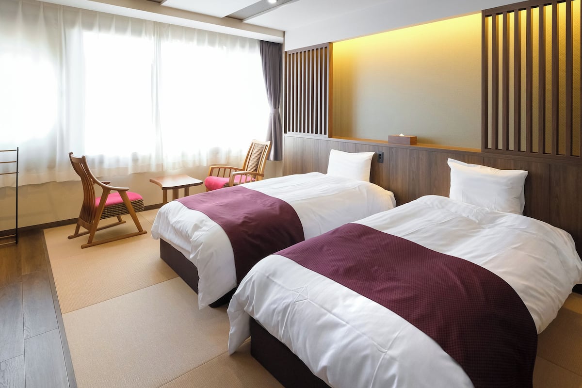 Une chambre d’hôtel avec deux lits, une petite table, des chaises et une grande fenêtre avec des rideaux.