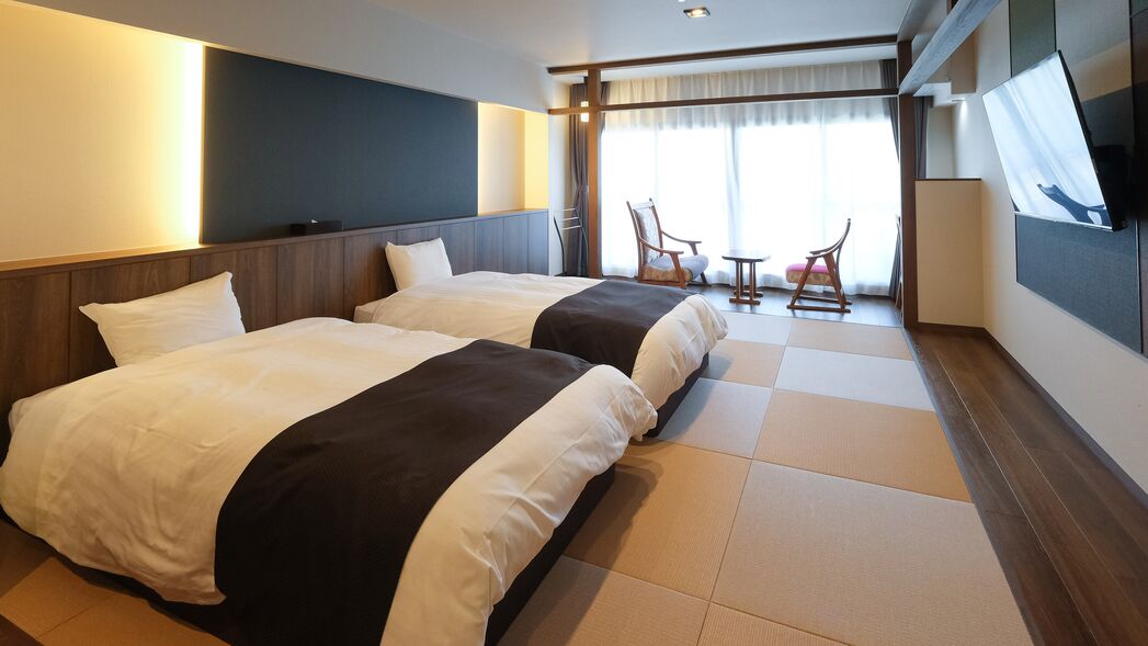 A hotel room with two beds, a TV, a seating area with chairs, and a large window.