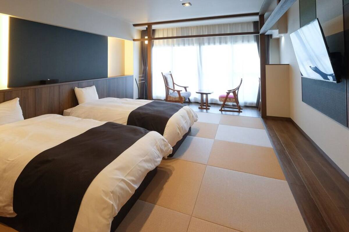 Une chambre d’hôtel avec deux lits, une télévision, un coin salon avec des fauteuils et une grande fenêtre.