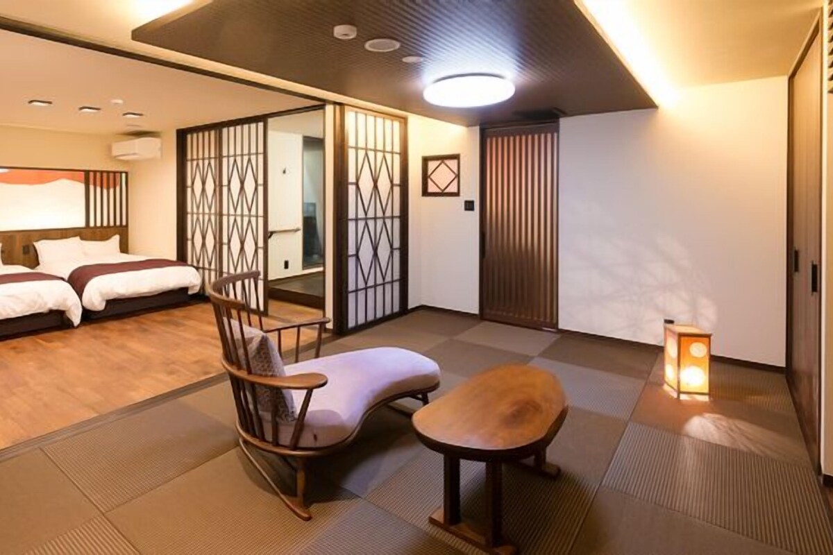 Una habitación tradicional japonesa con suelo de tatami, futón, mesa de madera baja, silla acolchada y puertas correderas shoji.