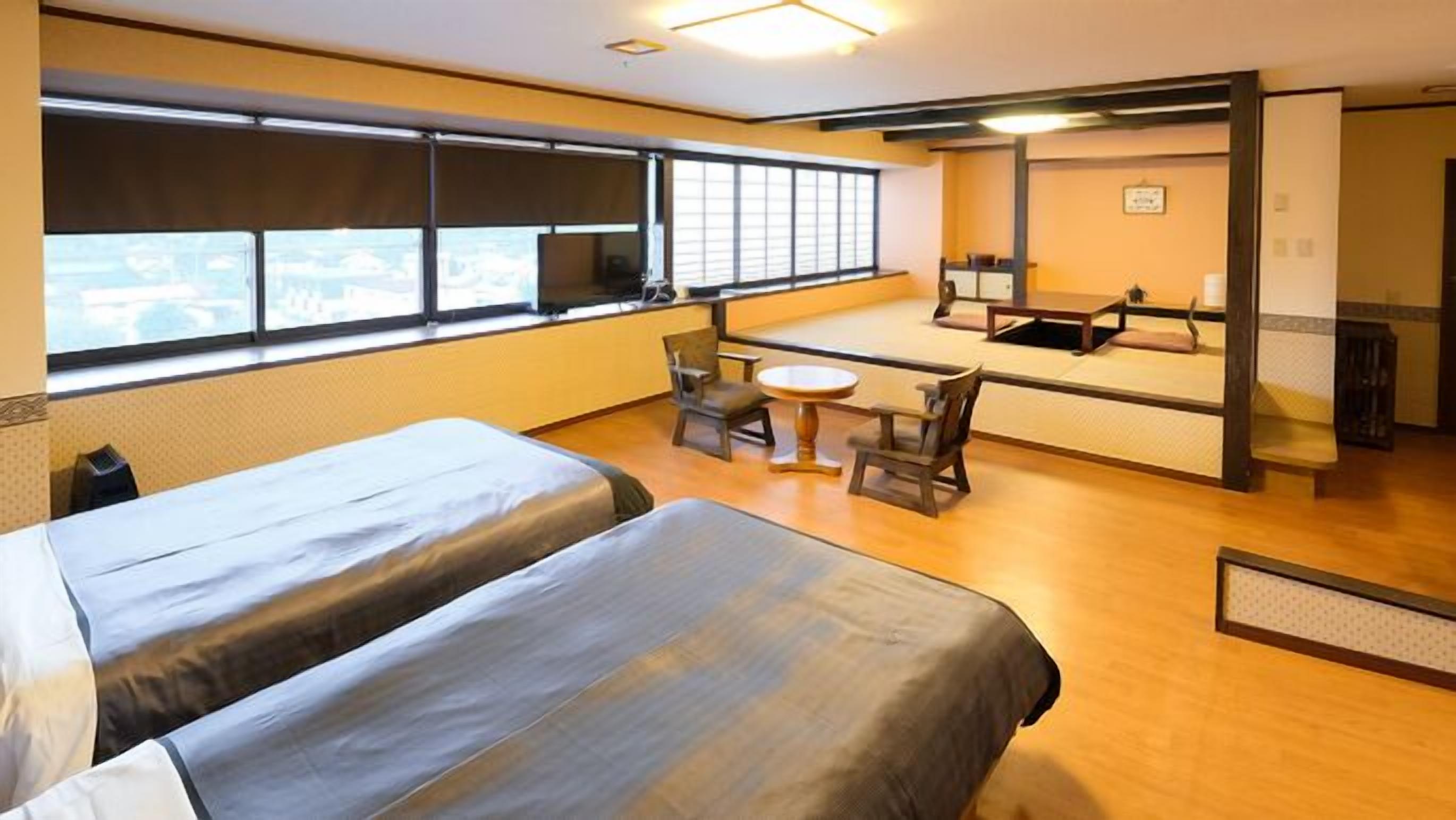 A hotel room with two beds, a small table, a chair, and a large window.