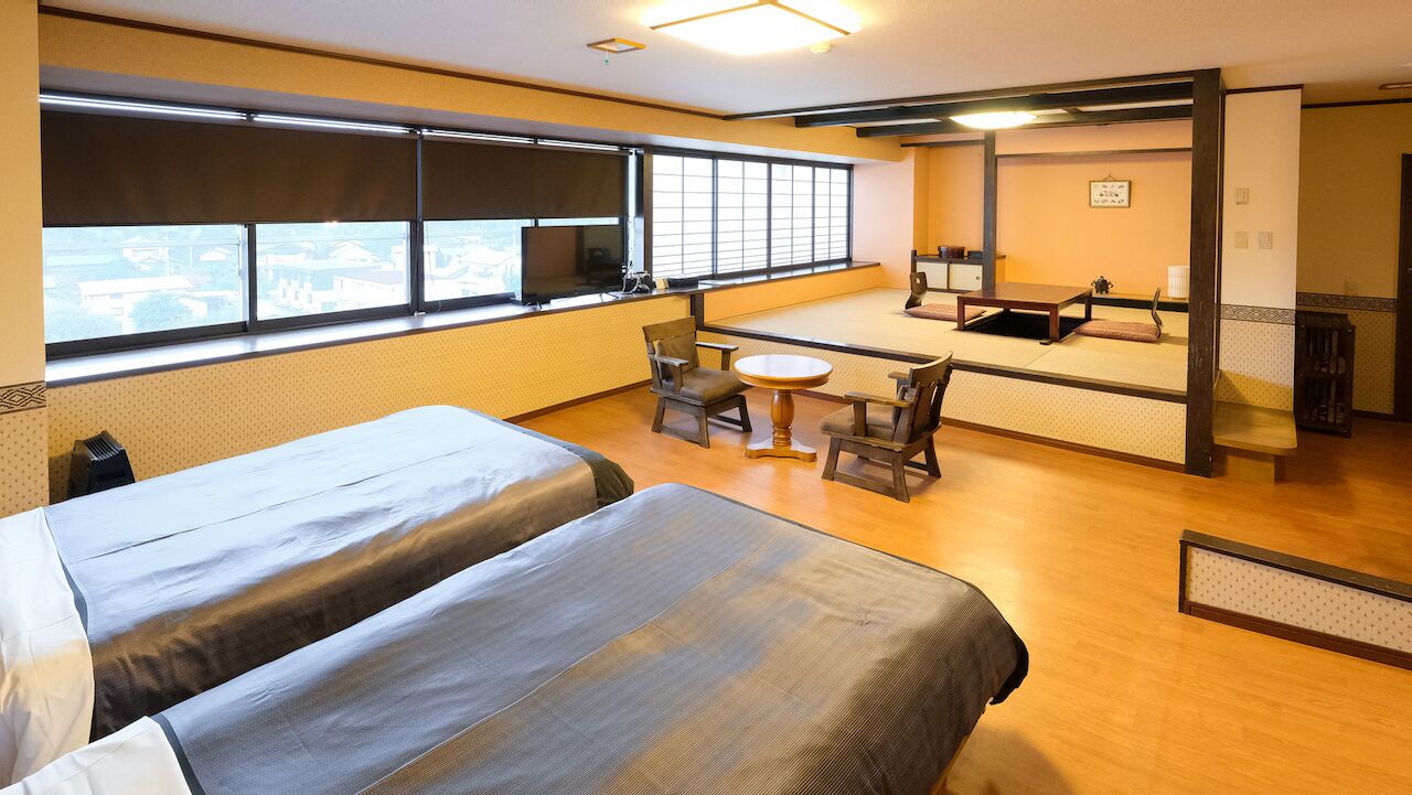 A hotel room with two beds, a small table, chairs, a TV, and a large window.