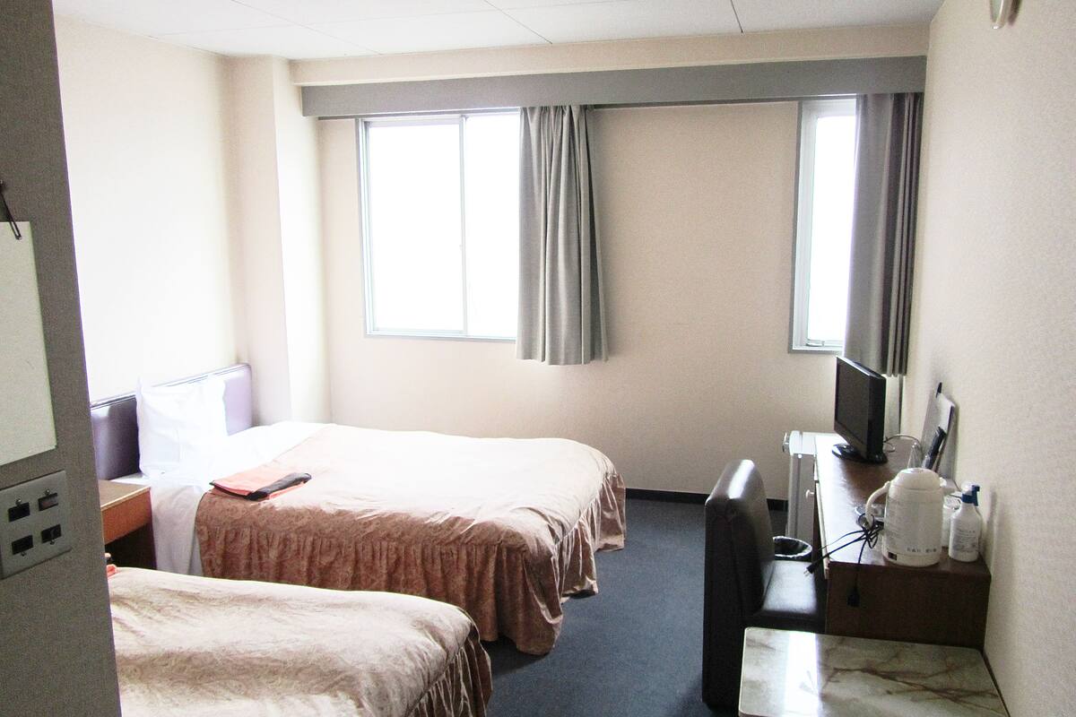 Une chambre d’hôtel avec deux lits, un bureau, une chaise, une télévision et une fenêtre avec des rideaux.