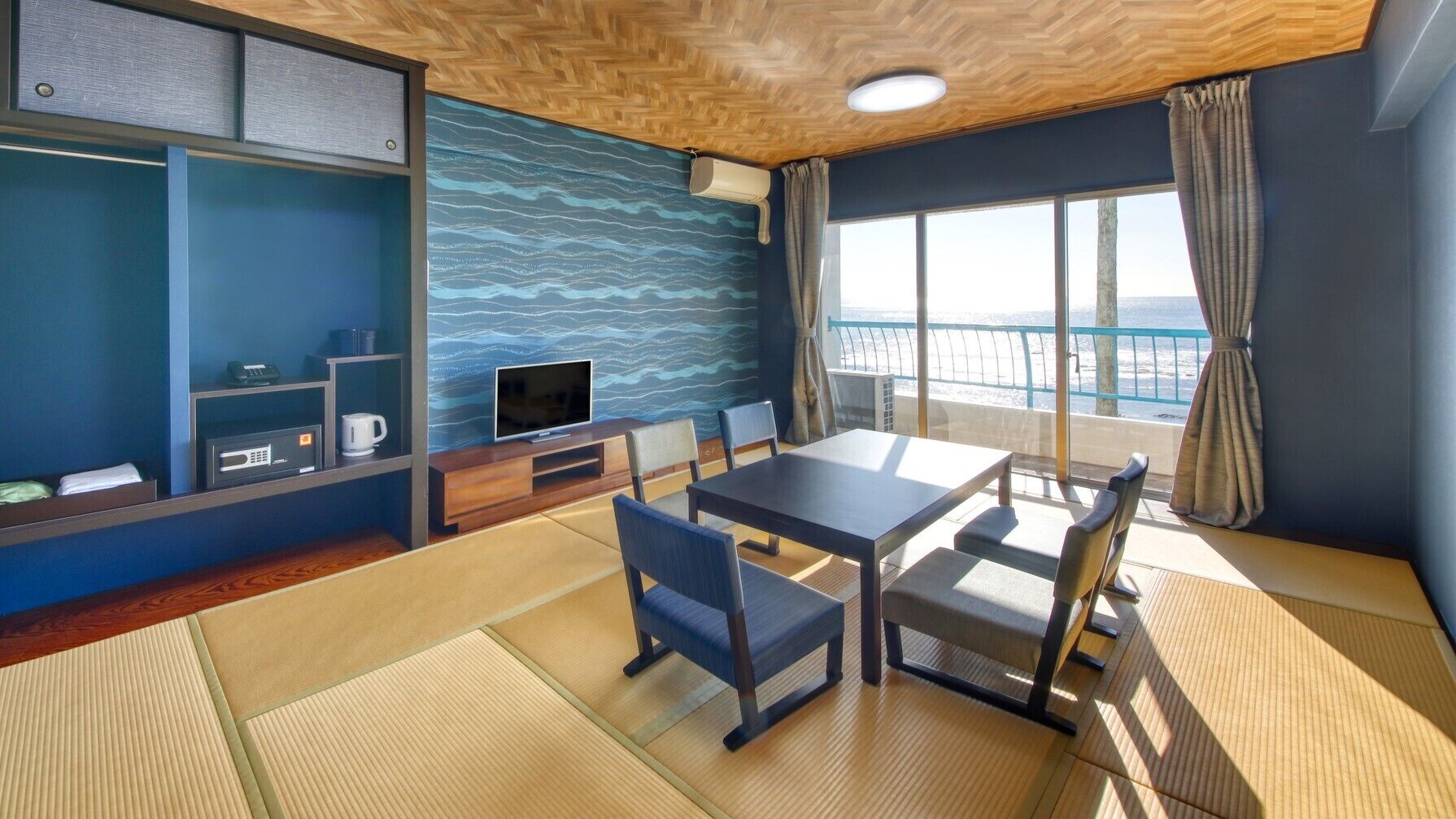 A room with a wooden ceiling, a television, a table with chairs, a sofa, a window with curtains, and a view of the sea.