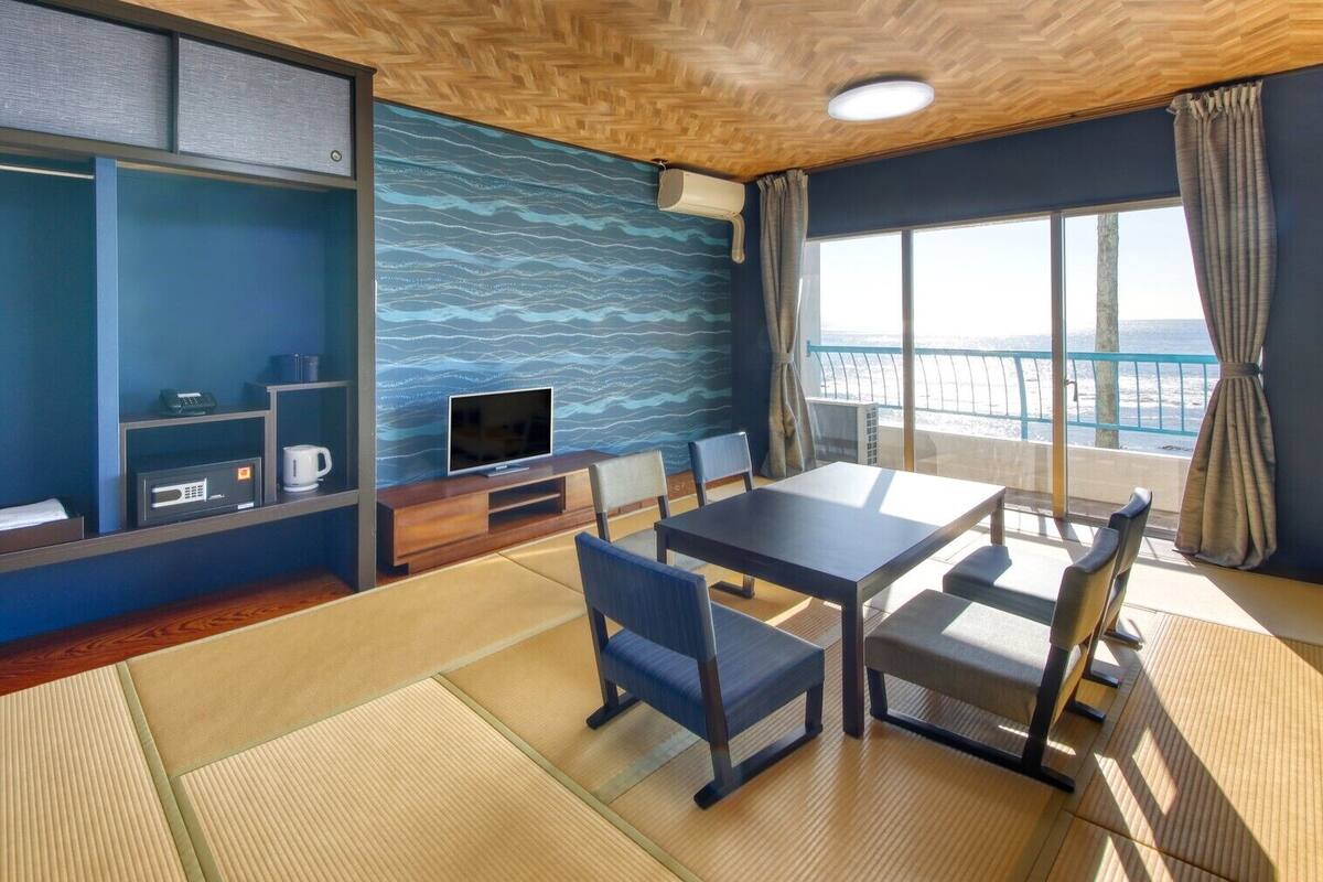A room with a wooden ceiling, a television, a table with chairs, a sofa, a window with curtains, and a view of the sea.