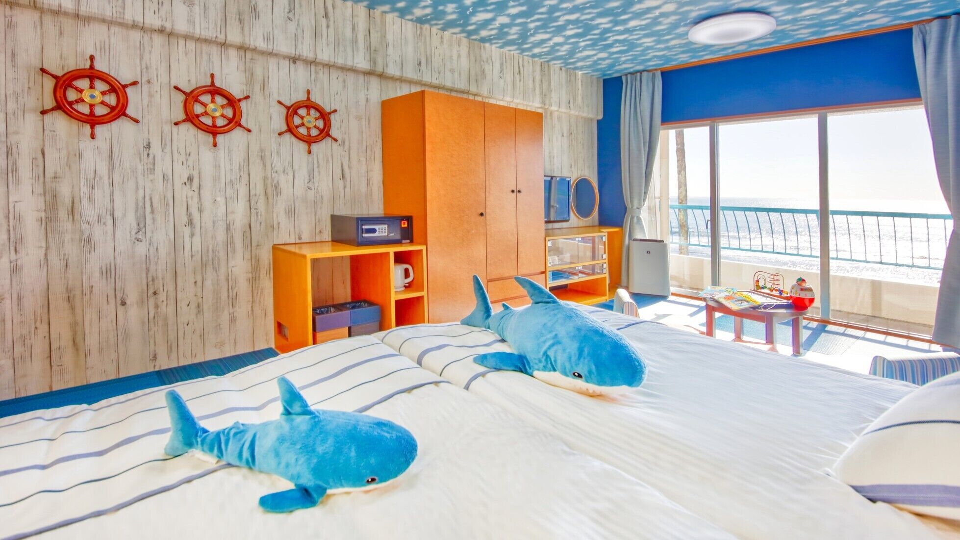 A room with a bed, two shark plush toys, a wooden wall with ship wheel decorations, a wooden cabinet, and a view of the sea through a large window.