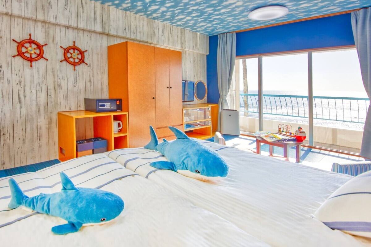 A room with a bed, two shark plush toys, a wooden wall with ship wheel decorations, a wooden cabinet, and a view of the sea through a large window.