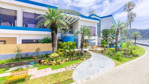A modern building with a blue and white facade, a landscaped garden with palm trees, and a curved driveway. - Kominato Miiri Onsen Hotel Green Plaza Kamogawa (Kamogawa)