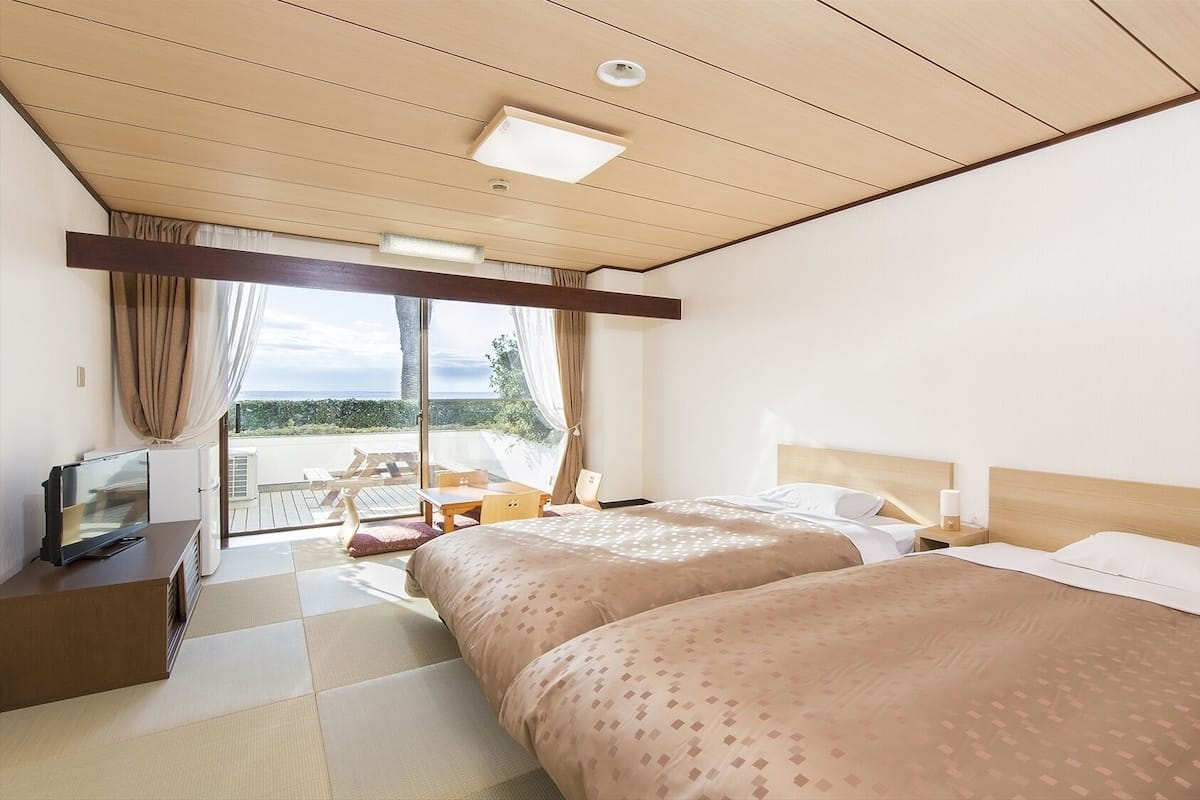 A hotel room with two beds, a TV, a desk, and a view of the sea.