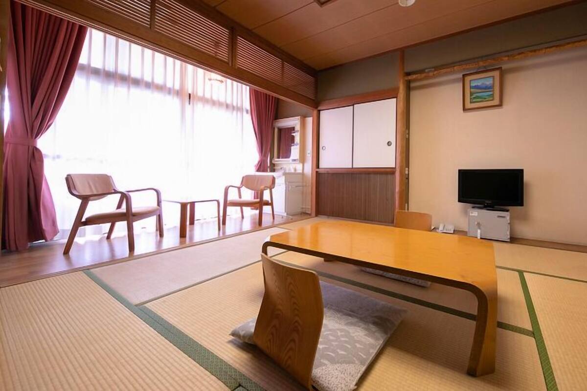 Une pièce de style japonais traditionnel, avec un sol recouvert de tatamis, une table basse, des chaises, un téléviseur et des portes coulissantes.