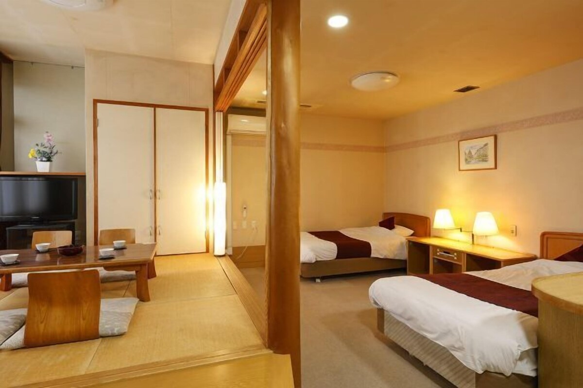 A hotel room with a traditional Japanese-style tatami mat floor, a wooden dining table with chairs, a television, and two beds with bedside tables and lamps.