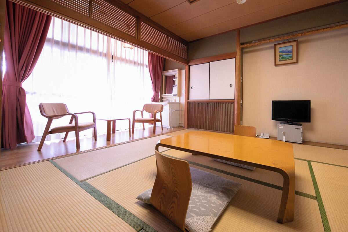 Une pièce de style japonais traditionnel, avec un sol recouvert de tatamis, une table basse, des chaises, un téléviseur et des portes coulissantes.