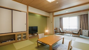 Um quarto japonês tradicional com tatames, uma mesa de madeira, cadeiras, uma televisão e uma porta deslizante para outro cômodo.