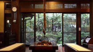 Een traditionele Japanse kamer met tatami matten, een waterkoker op een tafel, en uitzicht op een bos door grote ramen.