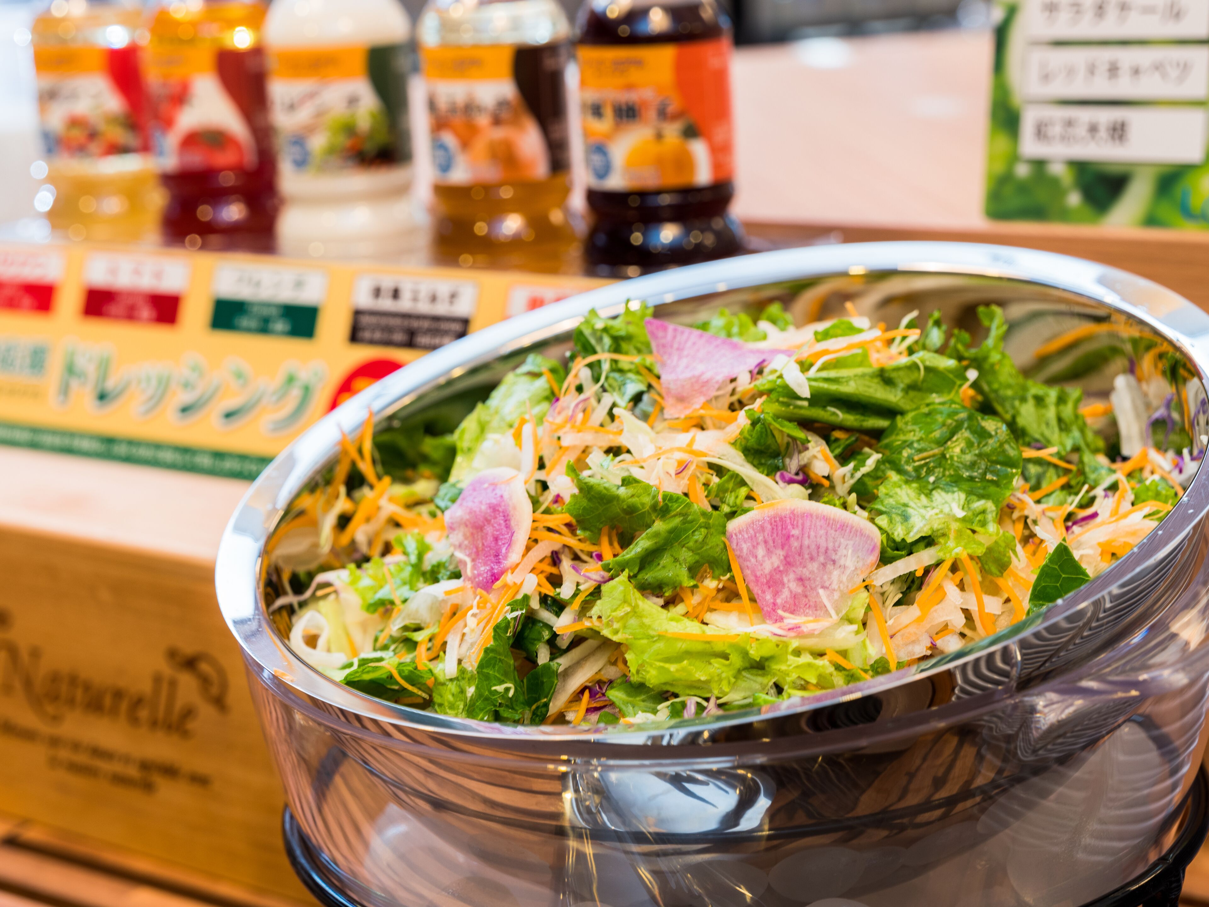 Un tazón de ensalada con diversos vegetales y zanahoria rallada, sobre una mesa con un letrero al fondo.