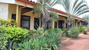 A building with a tiled roof, a covered porch, and a garden with various plants and a palm tree. - Dos Palmas Studio Apartments (Rio Segundo)
