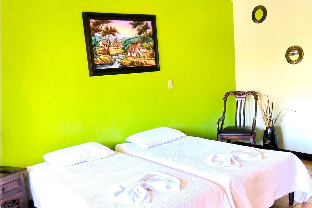 Una habitación con dos camas, un cuadro en la pared, una silla y un florero con ramas.