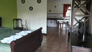 A room with a bed, a wooden bench, a small table with chairs, a kitchen area, and a window with curtains. - Dos Palmas Studio Apartments (Rio Segundo)