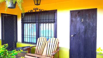 A wooden chair outside a building with a black door marked with the number 3, a window with bars, and a hanging plant.