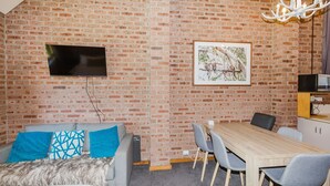 Ein Zimmer mit Backsteinwand, einem flachbildfernseher über einem grauen Sofa mit blauen Kissen, einem gerahmten Naturfoto, einem Holzesstisch mit grauen Stühlen und einem weißen Schrank mit einer Mikrowelle.