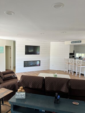 A modern living room with a fireplace, a television, and a kitchen area labeled "BEACH HOUSE". - Ocean Beach/Corneille Est, Fire Island Beach House (Ocean Beach (Cornielle Estates))