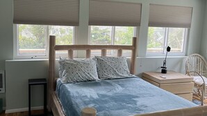 A bedroom with a wooden bed, a bedside table, a chair, and a window with blinds. - Ocean Beach/Corneille Est, Fire Island Beach House (Ocean Beach (Cornielle Estates))