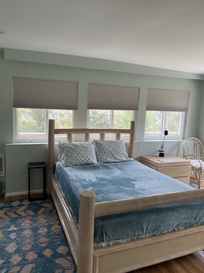 A bedroom with a wooden bed, a bedside table, a chair, and a window with blinds. - Ocean Beach/Corneille Est, Fire Island Beach House (Ocean Beach (Cornielle Estates))