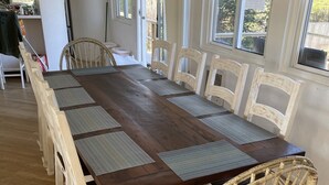 A large wooden dining table with woven chairs, surrounded by windows with blinds, in a bright room with hardwood flooring. - Ocean Beach/Corneille Est, Fire Island Beach House (Ocean Beach (Cornielle Estates))