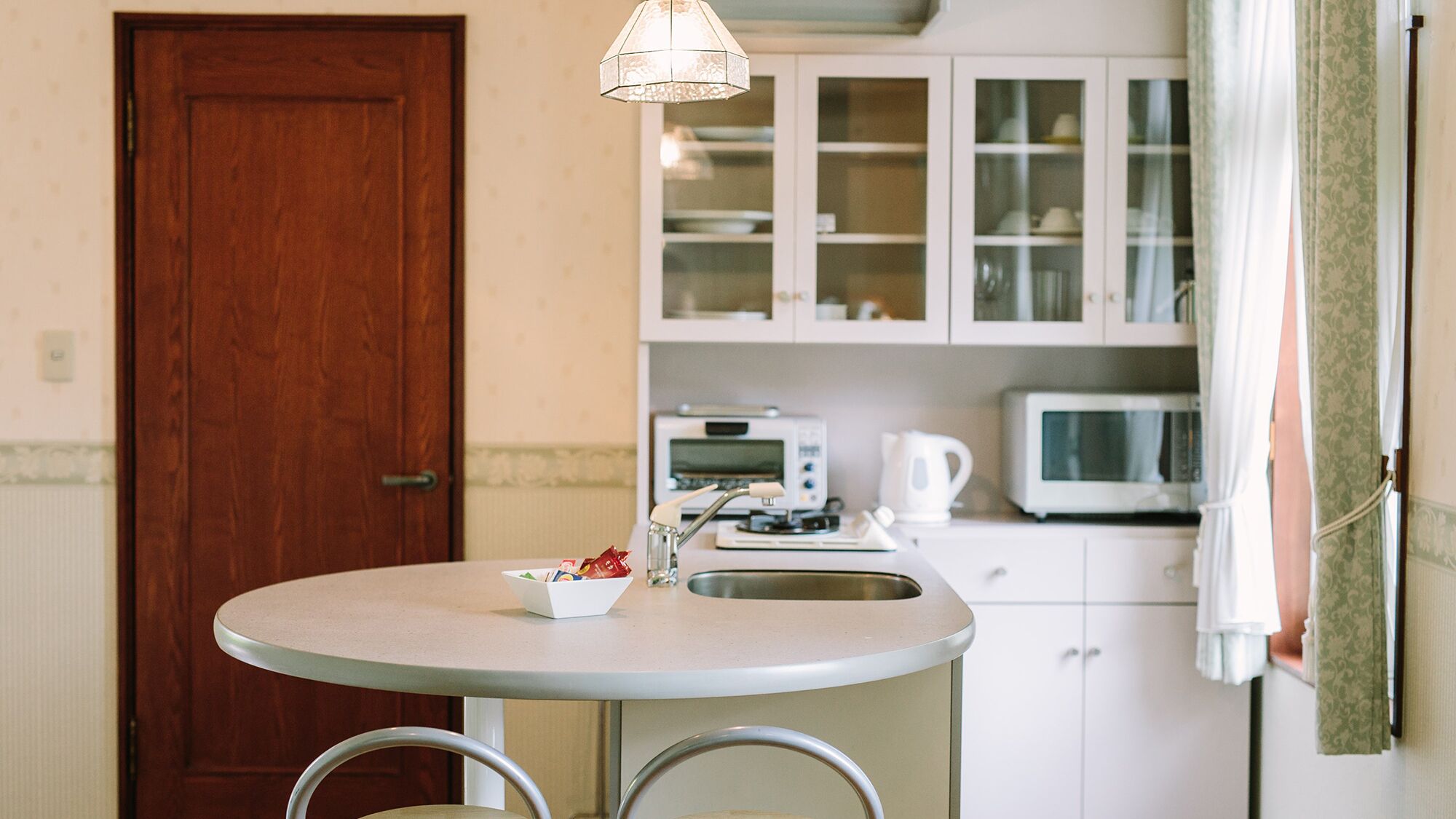 Een kleine keuken met een ronde tafel, twee stoelen, een magnetron, een waterkoker en een kast met glazen deuren.
