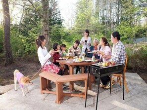 Un groupe de personnes est assis à une table en bois, entourée de bancs, et prend son repas en plein air, au milieu des arbres.