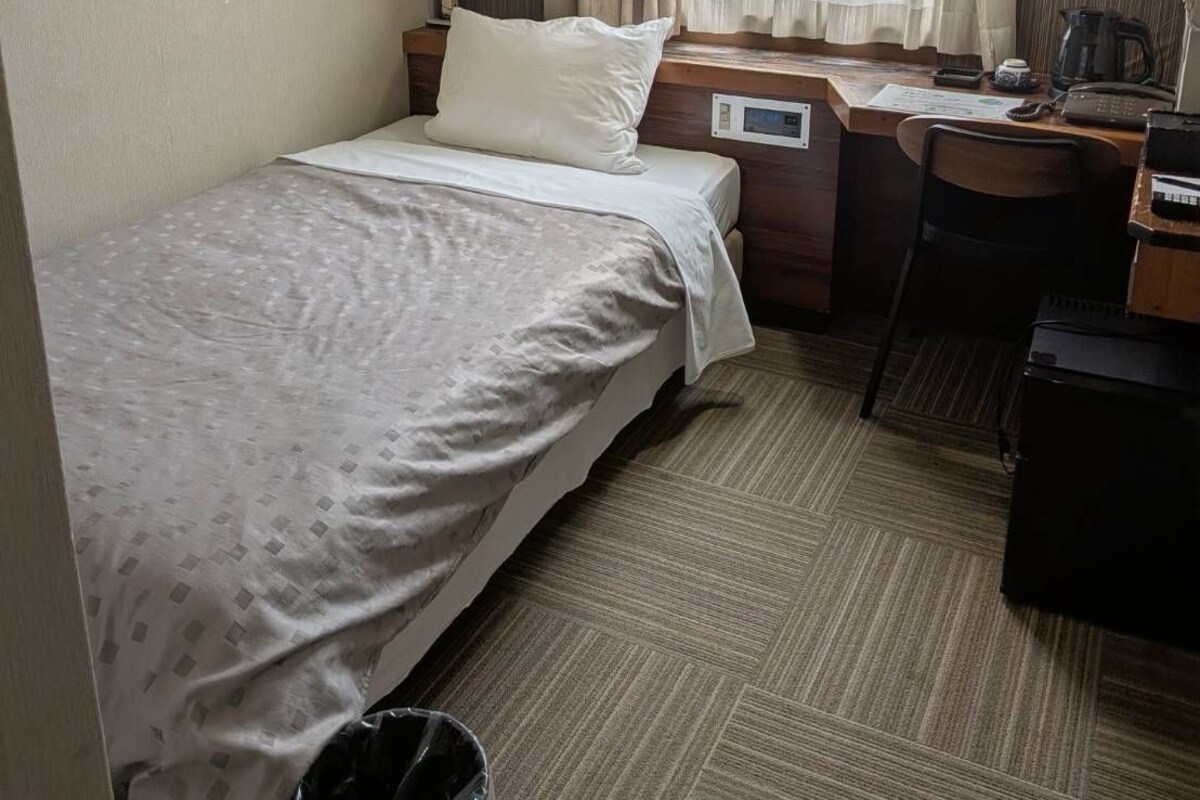 Une chambre d’hôtel avec un lit, un bureau, une chaise, une poubelle et une fenêtre avec des rideaux.