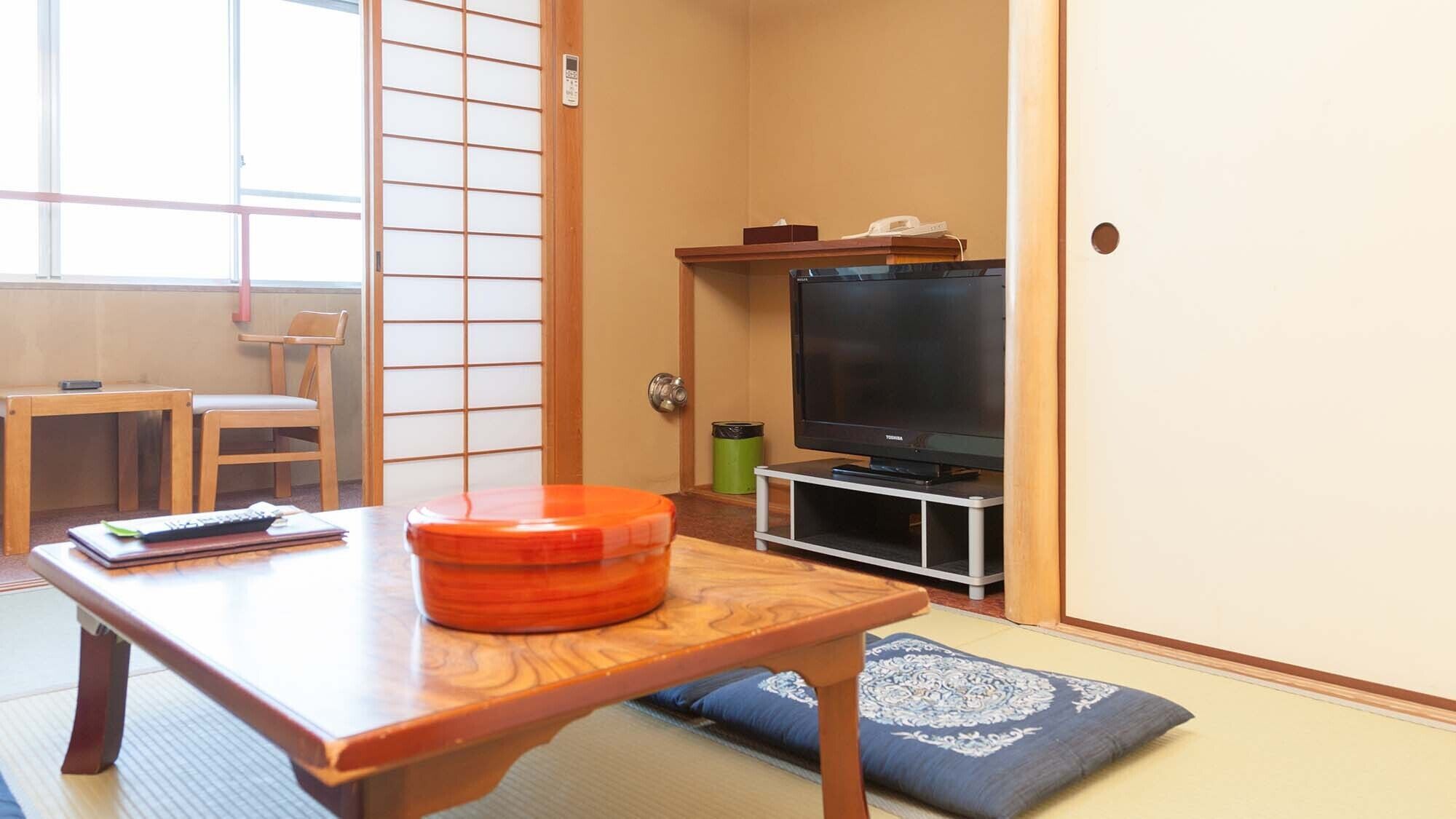 Habitació japonesa tradicional amb sòls de tatami, un televisor sobre un suport, una taula de fusta amb coixí i una porta corredissa.