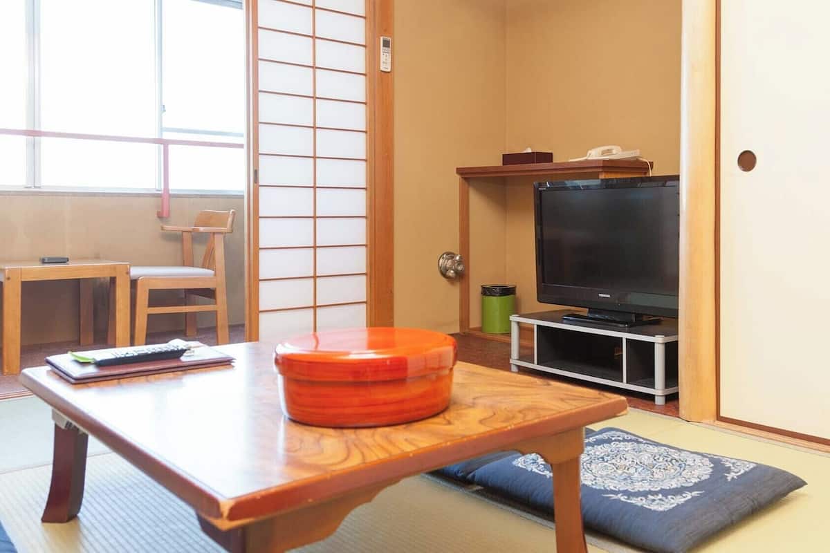 A traditional Japanese room with tatami flooring, a television on a stand, a wooden table, and a sliding door.
