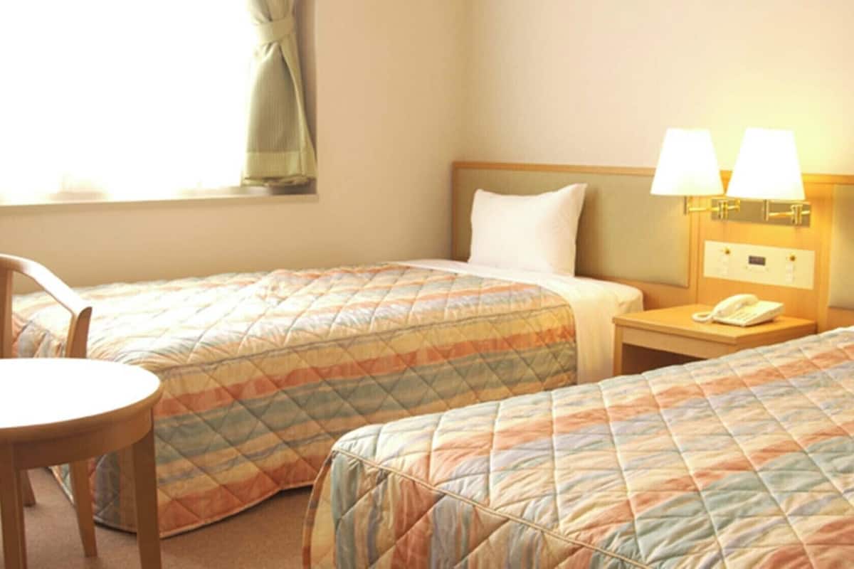 A hotel room with two beds, a chair, a nightstand with a lamp, and a window with curtains.