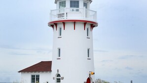 Un phare blanc avec une porte rouge et un balcon, entouré d’herbe et bordé d’une route.