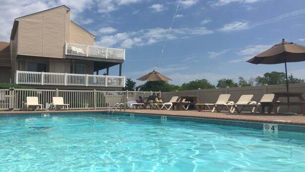 Une piscine avec des chaises longues et des parasols, un bâtiment doté d’un balcon et un ciel dégagé.