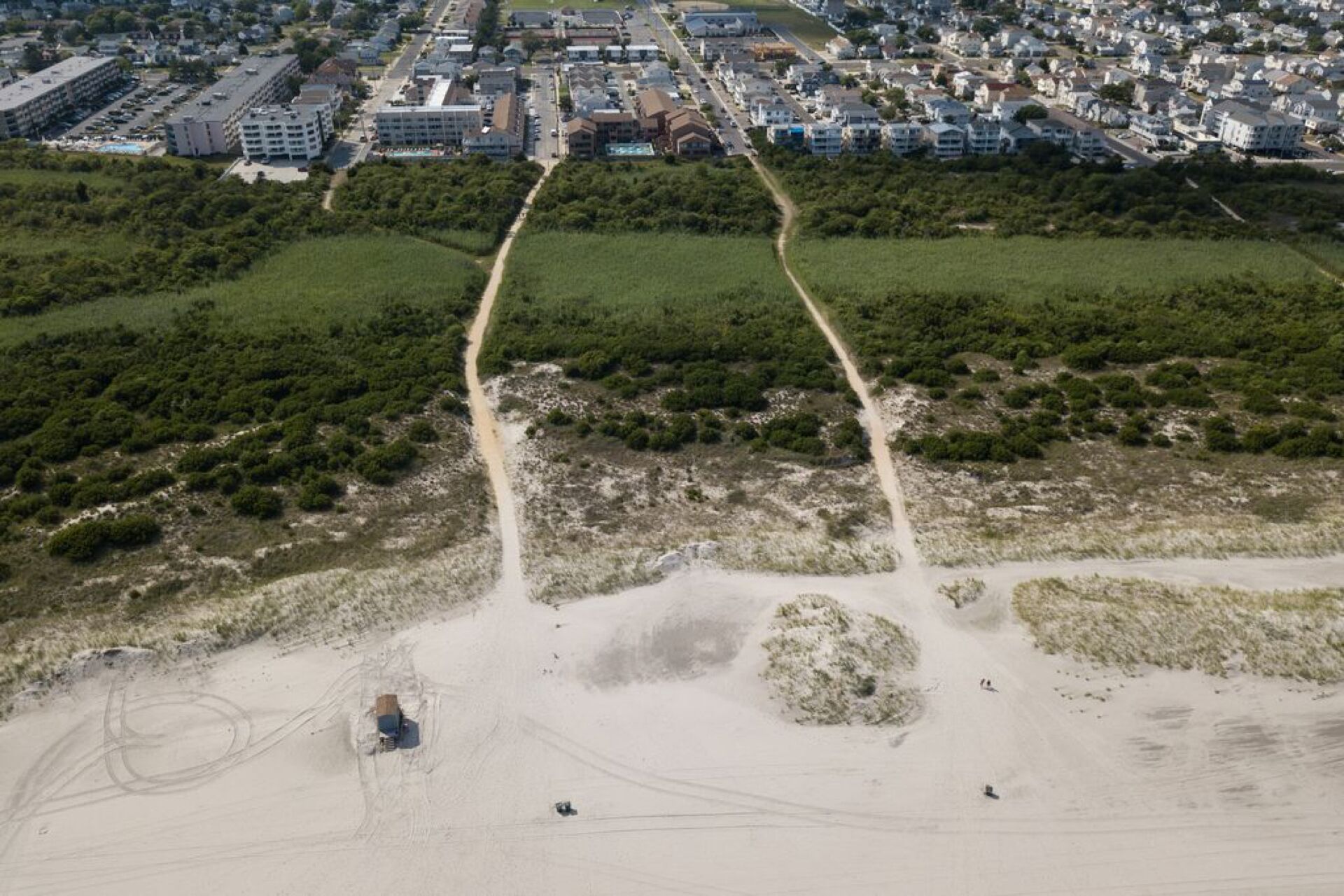 An aerial view of a coastal area with a sandy beach, a dirt path leading to a small structure, and a densely built urban area in the background.