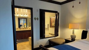 A hotel room with a bed, a nightstand, a lamp, a mirror, and an open door leading to a bathroom with a sink and mirror. - Ohana Manor, Clean and Comfortable, Sleeps 6 (Kapaʻa)