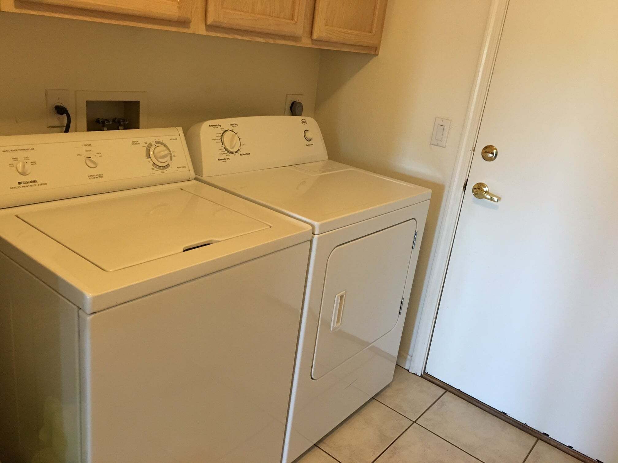 A laundry room with two front-loading washing machines and a dryer, wooden cabinets above, and a white door with a keyhole.
