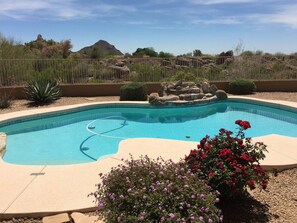A swimming pool with a rock feature, surrounded by a fence, with desert vegetation and a mountain in the background.