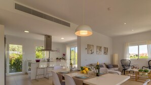 Ampla sala de estar com mesa de jantar, cadeiras e sofá. Há uma cozinha com fogão e pia. O quarto tem grandes janelas com cortinas.