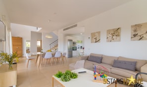 A modern living room with a sofa, dining table, and chairs. There is a kitchen area in the background with a refrigerator and microwave. - Brisa de Mar 1, Villa 5StarsHome Mallorca (Alcúdia)