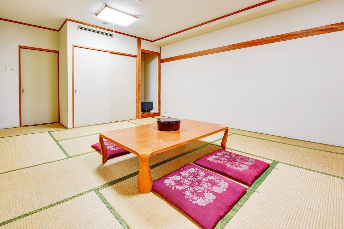 Ein Raum mit Tatami-Matten, einem Holztisch, Kissen, einer Schiebetür und einer Deckenleuchte.