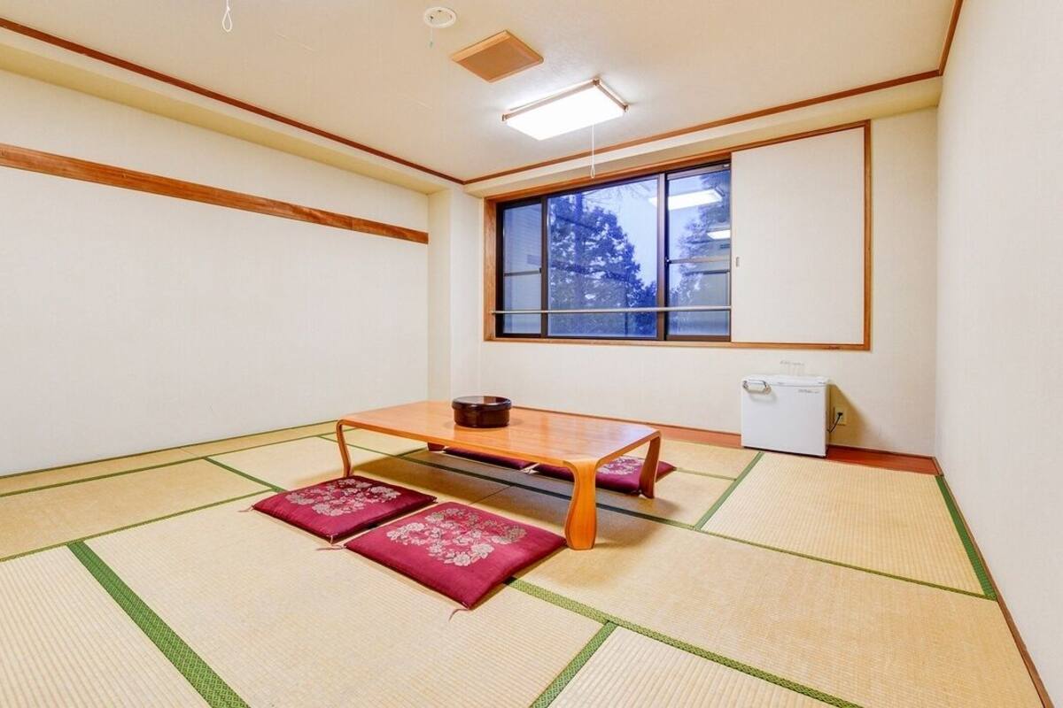 Ein Raum mit Tatami-Matten, einem niedrigen Tisch, Kissen, einem Fenster und einem kleinen Kühlschrank.