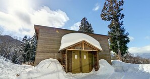 Eine Holzhütte mit verschneitem Dach und zwei Türen, umgeben von Schnee und Bäumen.